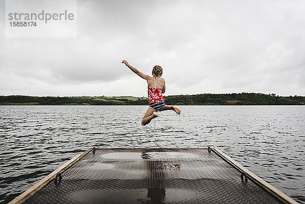 Kleines Mädchen springt an einem bewölkten Sommertag vom Dock in den See