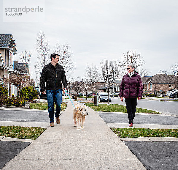 Mann und ältere Dame gehen mit dem Hund auf dem Bürgersteig eines Vorstadtviertels spazieren.