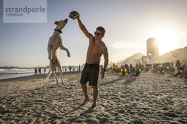 Mann spielt mit springendem Hund und Kokosnuss am Strand von Copacabana