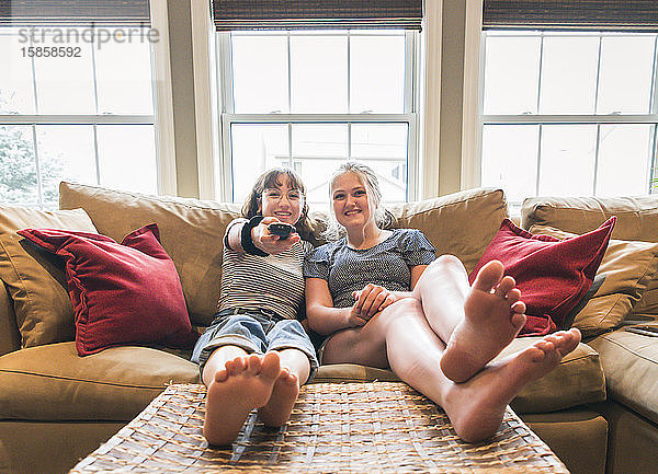 Zwei Teenagermädchen sitzen mit erhobenen Füßen auf einer Couch und schauen fern.