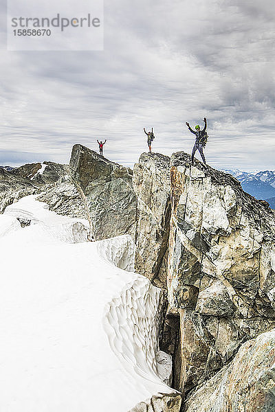 Drei Bergsteiger stehen mit erhobenen Armen auf einem felsigen Gipfel