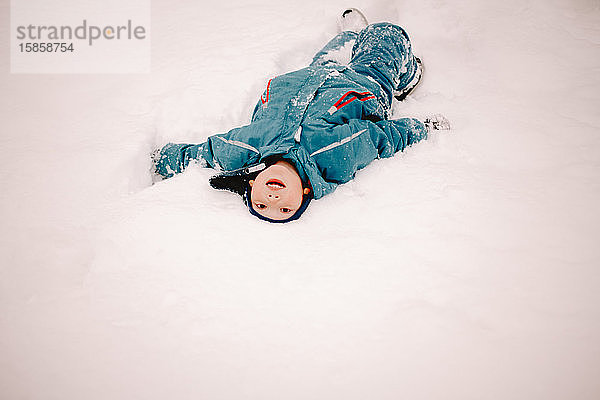 Fröhlicher Junge mit offenem Mund  der im Winter im Schnee liegt