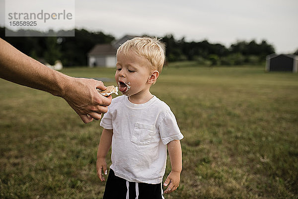 Kleinkind öffnet den Mund  um im Hinterhof s'more von der Hand des Vaters zu essen
