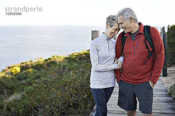 Glückliches älteres Ehepaar mit Arm in Arm auf der Strandpromenade am Meer gegen den Himmel stehend