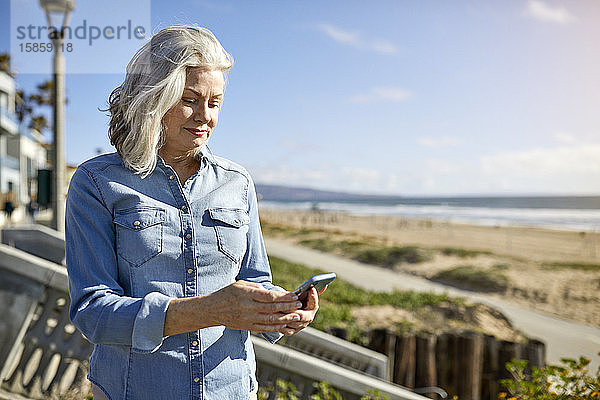 Ältere Frau benutzt ein Smartphone  während sie am Strand von Manhattan an einem sonnigen Tag gegen den Himmel steht