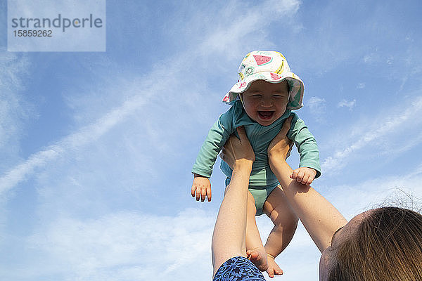 Mutter hält kleines Mädchen mit beiden Armen gegen einen blauen Himmel in die Luft