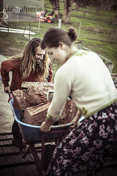 junges Paar Mann und Frau stapeln Holz in Schubkarre auf dem Bauernhof
