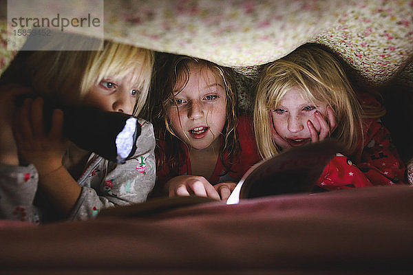 Drei kleine Mädchen lesen unter einer Decke mit Taschenlampe ein Buch