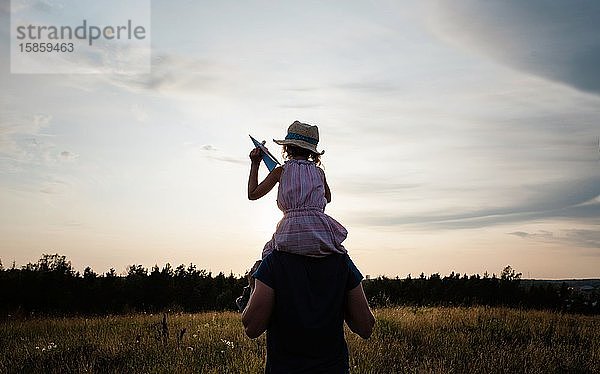 Vater  der seine Tochter auf den Schultern trägt und bei Sonnenuntergang spazieren geht