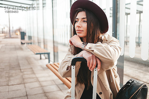 Nachdenkliches stilvolles Mädchen in Mantel und Hut sitzt an einer Haltestelle und wartet auf den Bus