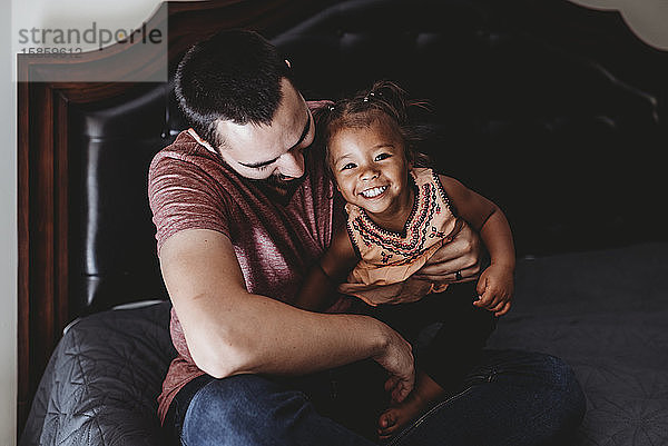 Dunkelhaariger Vater sitzt mit lächelndem Mischlingsvater im Bett  2 Jahre alt
