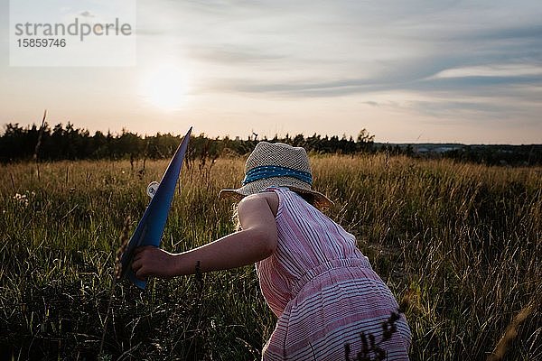 Mädchen  das im Sommer bei Sonnenuntergang auf einer Wiese mit Papierflugzeugen spielt