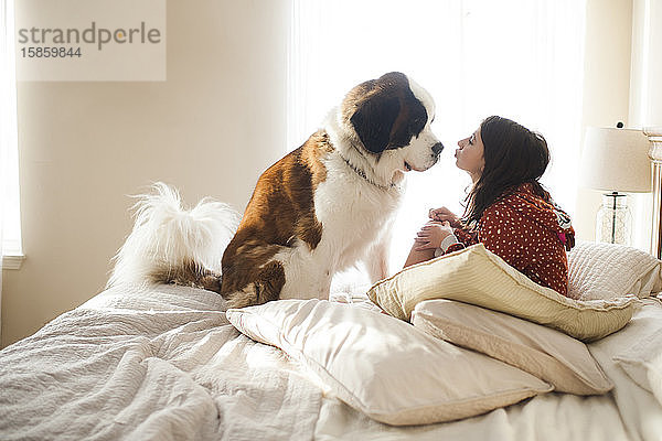 Junges Mädchen sitzt auf dem Bett mit großem Hund  der ihn gleich auf die Nase küssen wird