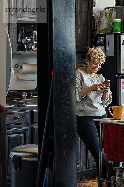 Lächelnde mittelgroße erwachsene Frau  die zu Hause in der Küche steht und Textnachrichten auf einem Smartphone verschickt