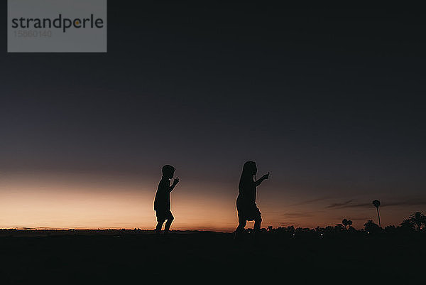 Silhouette eines Geschwisterpaares  das bei Sonnenuntergang am Strand spazieren geht