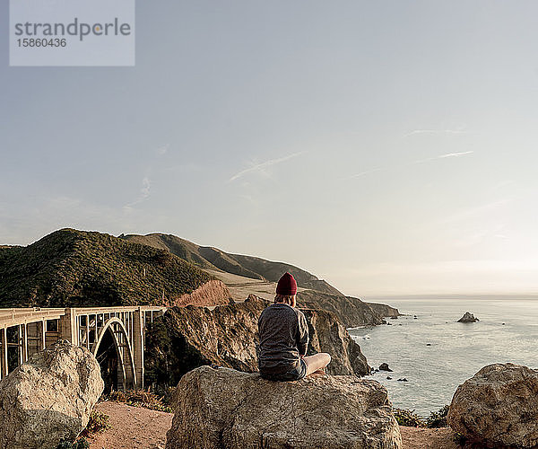 Frau sitzt auf einem Felsen mit Blick auf die Küste von Big Sur und Bixby Br