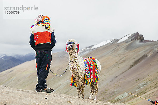 Ein einheimischer Peruaner steht in Peru in der Nähe eines Lamas mit Sonnenbrille