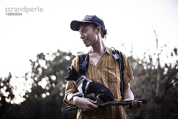 Natürliche androgyne Person mit Mütze hält winzigen Hund auf Holz im Parklicht