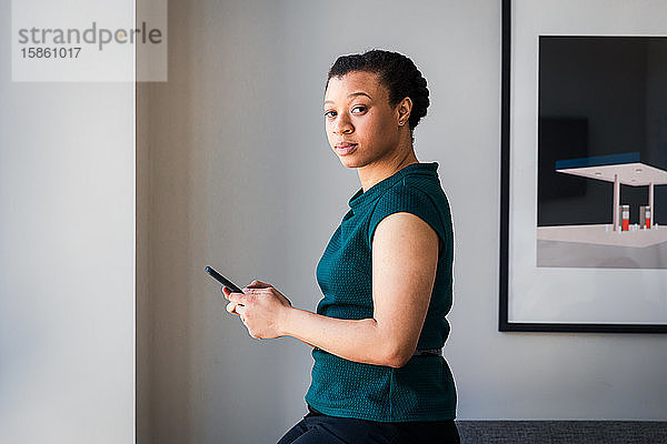 Seitenansicht der Textnachrichten einer Geschäftsfrau auf einem Smartphone  während sie im Büro an der Wand steht