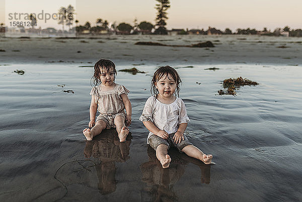 Frontansicht von Kleinkind-Schwestern  die am Strand im Wasser sitzen