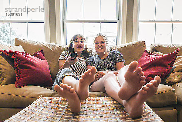 Zwei Mädchen im Teenageralter sitzen auf einer Couch und sehen mit erhobenen Füßen fern.