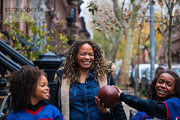 Glückliches Mädchen hält American Football in der Hand  während es Schwester und Mutter auf einem Fußweg in der Stadt zur Seite steht