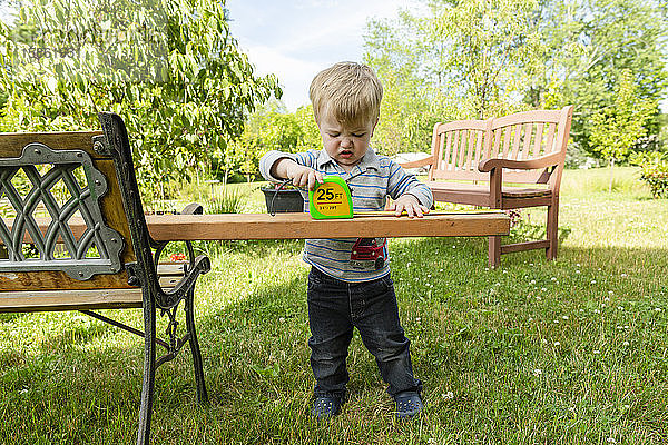 Kleinkind Junge sieht frustriert aus  während er im Freien ein Holzbrett misst