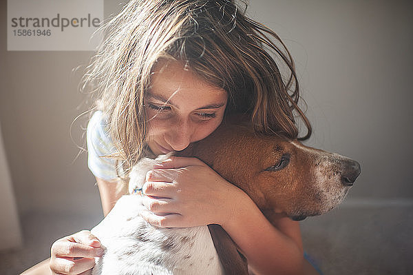 Happy Tween Girl umarmt ihren Basset-Hundhund in der Nähe eines Fensters