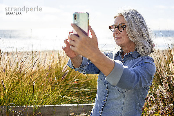 Ältere Frau  die bei sonnigem Wetter am Strand von Manhattan Selfie nimmt  während sie am Strand von Manhattan steht