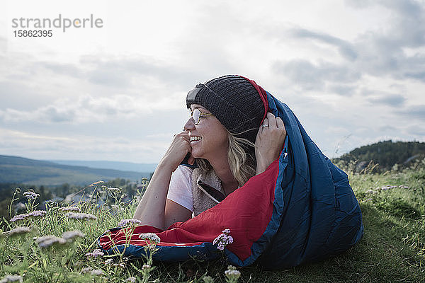 Frau liegt im Schlafsack auf einem Berg und schaut lächelnd auf die Aussicht
