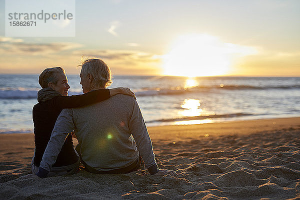 Rückansicht eines älteren Ehepaares  das bei Sonnenuntergang am Strand im Sand sitzt