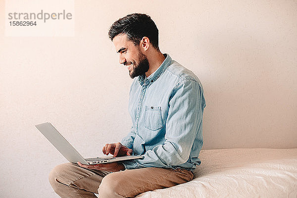 Glücklicher Mann benutzt Laptop-Computer  während er zu Hause an der Wand sitzt