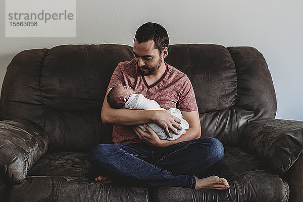Barfüssiger bärtiger Papa auf Ledercouch  der ein gewickeltes Neugeborenes hält