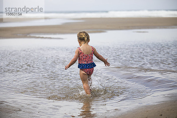 Rückansicht eines jungen Mädchens  das durch ein Gezeitenbecken am Strand läuft.