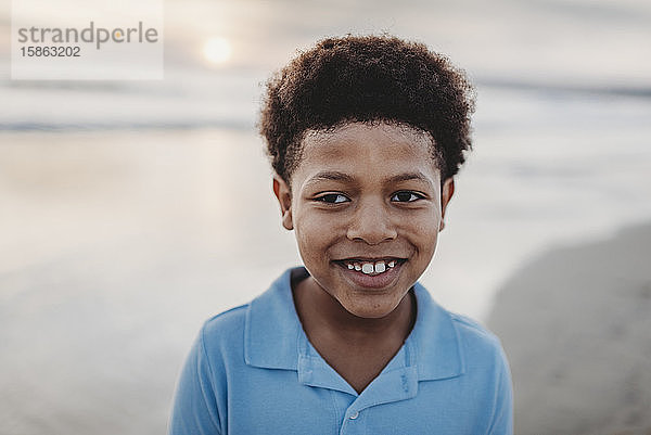 Porträt eines jungen Jungen im Schulalter  der bei Sonnenuntergang am Strand lächelt