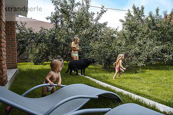Kleines Mädchen spielt mit Wasser im Hof eines Hauses mit einem schwarzen Hund