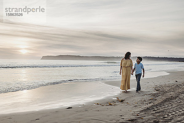 Junge Mutter mit Kleid und Sohn gehen bei bewölktem Sonnenuntergang am Strand