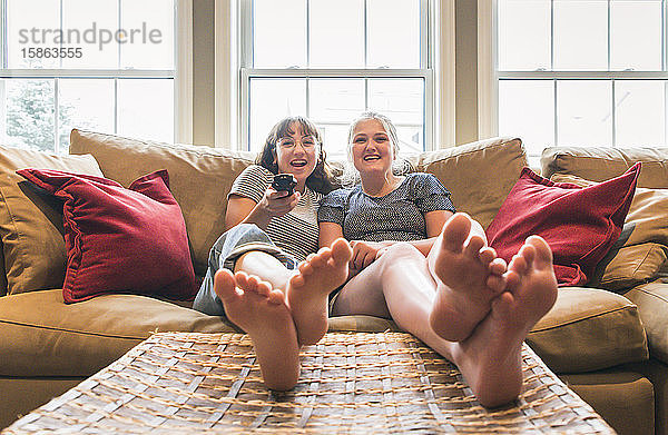 Zwei Teenagermädchen sitzen mit erhobenen Füßen auf einer Couch und schauen fern.