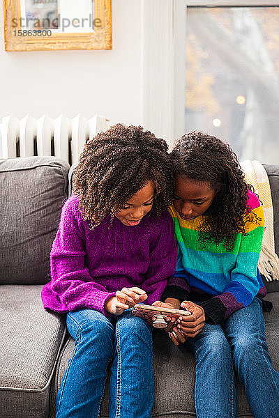 Schwestern schauen auf das Smartphone  während sie auf dem Sofa im Wohnzimmer sitzen