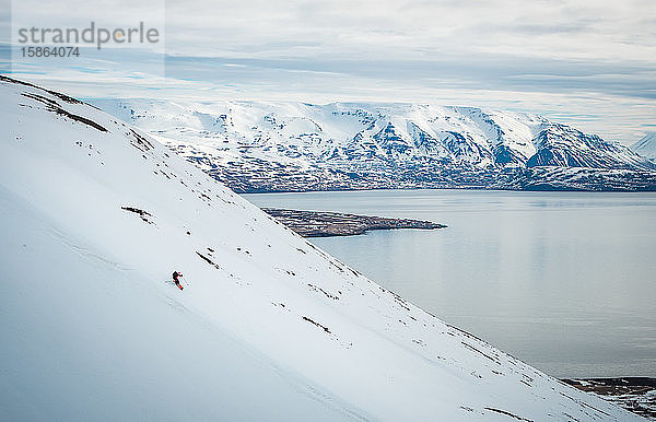 Ein Mann fährt auf Skiern einen Berg hinunter mit schneebedeckten Bergen und dem Meer dahinter