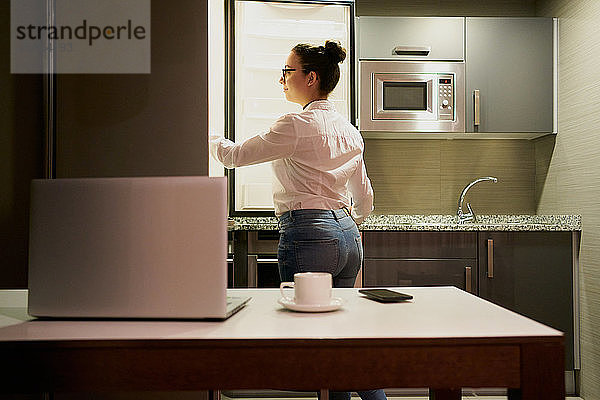 Eine junge Frau  die mit einem Laptop arbeitet  schaut in den Kühlschrank