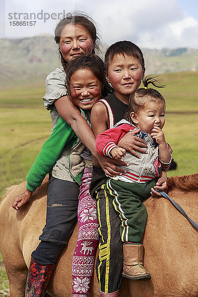 Porträt von vier lächelnden und glücklichen Nomadengeschwistern auf ihrem zahmen Pferd im Sommernomadenlager  Khujirt  Uvurkhangai  Zentralmongolei  Zentralasien  Asien