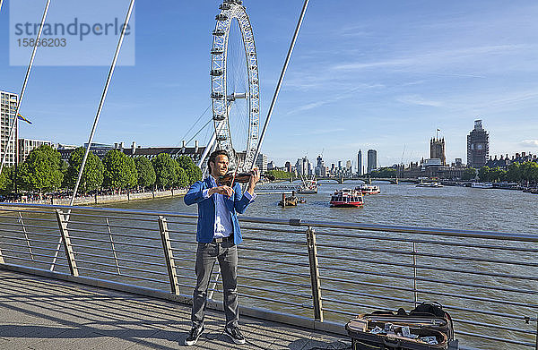Geige spielender Straßenmusiker auf einer Brücke über die Themse mit Booten und dem London Eye im Hintergrund; London  England
