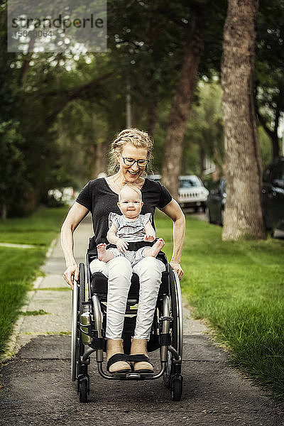 Eine querschnittsgelähmte Mutter  die ihr Baby auf dem Schoß trägt  während sie einen Rollstuhl benutzt  an einem warmen Sommernachmittag im Freien: Edmonton  Alberta  Kanada