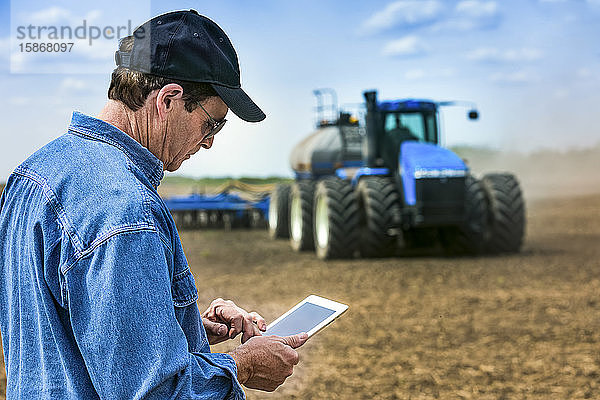 Landwirt  der ein Tablet benutzt  während er auf einem landwirtschaftlichen Feld steht und ein Traktor und Geräte das Feld besäen; Alberta  Kanada