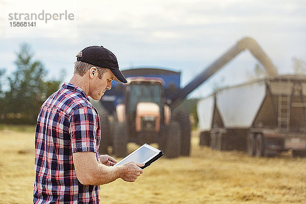 Ein Landwirt nutzt sein Tablet  um die Weizenernte zu steuern  während ein Mähdrescher eine volle Ladung Getreide auf einen Getreidewagen umlädt: Alcomdale  Alberta  Kanada