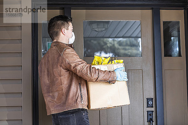 Ein junger Mann steht mit einer Kiste voller Lebensmittel vor der Tür eines Hauses während der Weltpandemie Covid-19; Edmonton  Alberta  Kanada