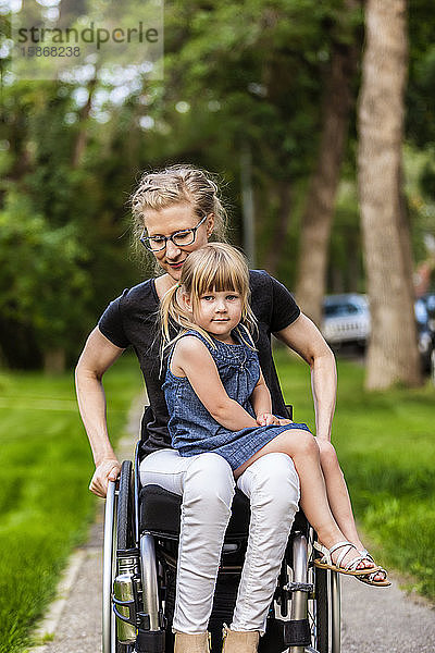 Eine querschnittsgelähmte Mutter hält ihr kleines Mädchen auf dem Schoß  während sie es an einem warmen Sommernachmittag in ihrem Rollstuhl die Straße hinunterfährt: Edmonton  Alberta  Kanada.