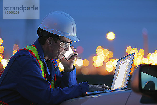 Mann arbeitet an einem Laptop und einem Smartphone mit einer Ölraffinerie im Hintergrund; Alberta  Kanada