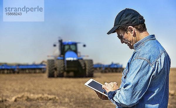 Landwirt  der ein Tablet benutzt  während er auf einem landwirtschaftlichen Feld steht und ein Traktor und Geräte das Feld besäen; Alberta  Kanada